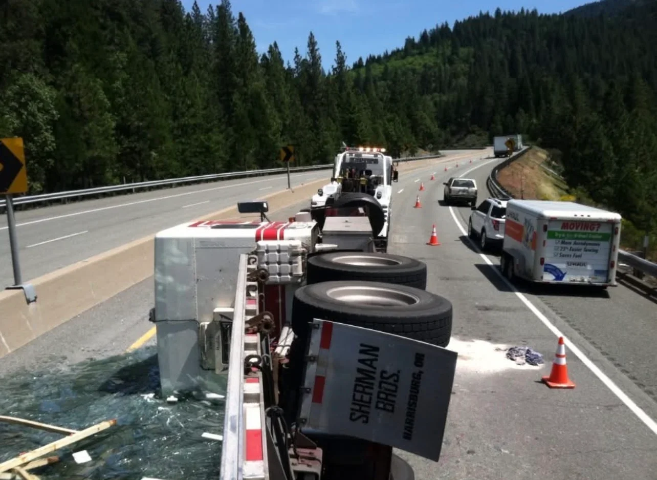 An overturned semi-truck blocking a mountain highway with police, tow trucks, and several cars present. Orange traffic cones are set along the road, and the truck's cargo spilled onto the road.