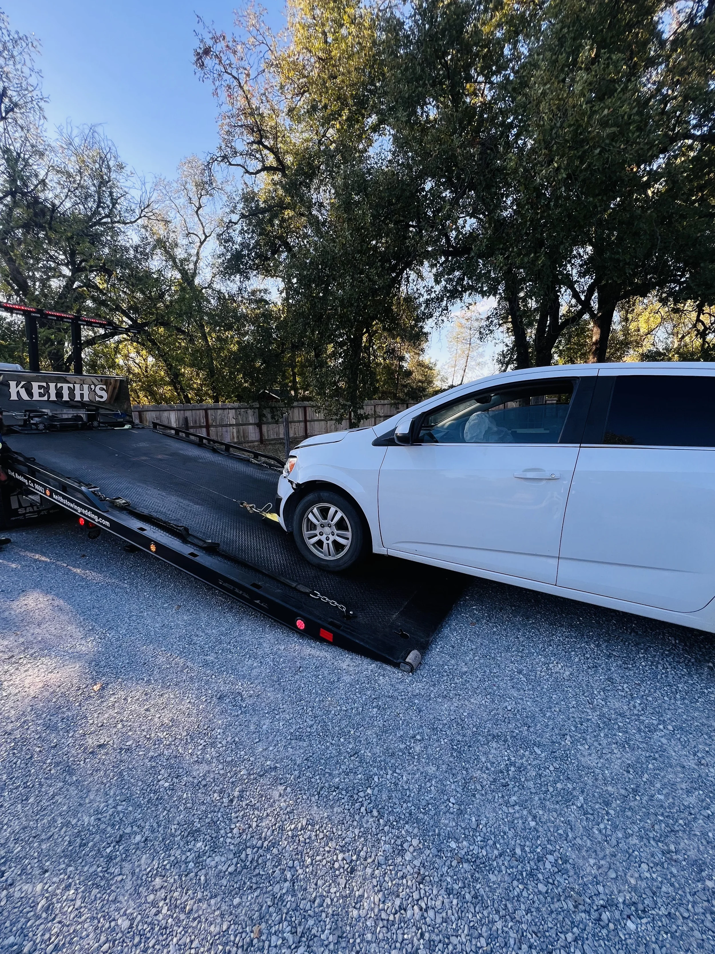 White car being loaded onto a flatbed tow truck on a gravel surface with trees and a wooden fence in the background.