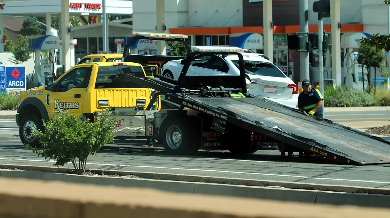 Tow truck from Keith's Towing is hauling a white car on a flatbed at a gas station with ARCO pumps. A person is standing beside the truck, and several vehicles are visible in the background.
