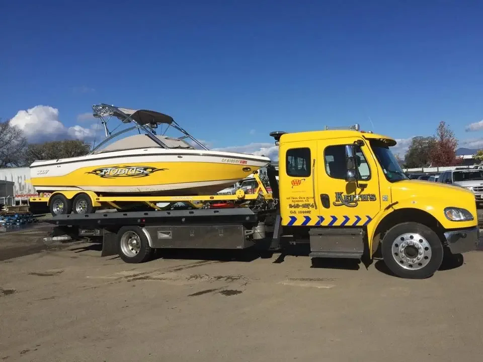 Yellow tow truck carrying a white and yellow speed boat with black and yellow graphics on a dirt lot under a blue sky with some clouds.