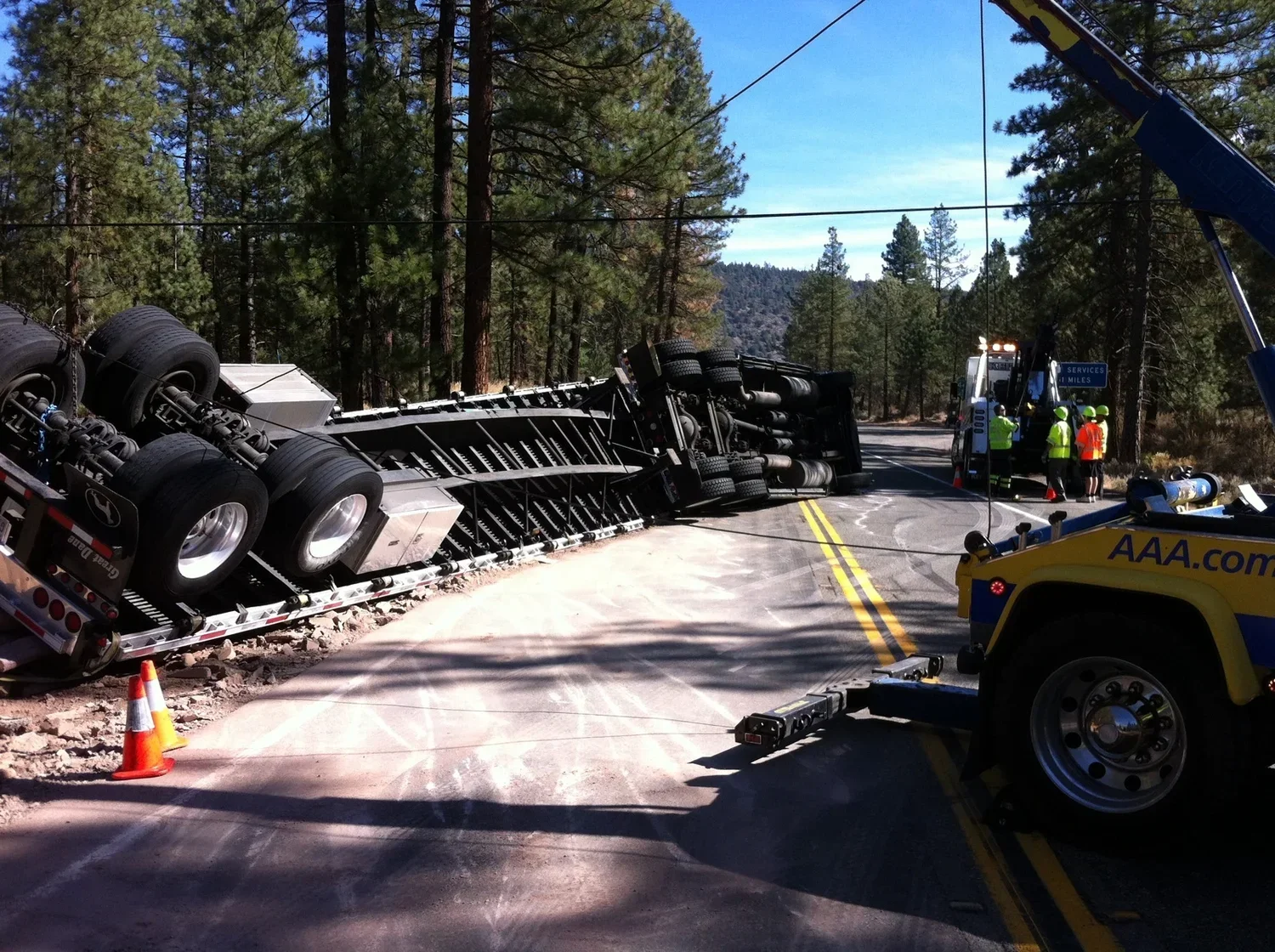 A semi-truck has overturned on a two-lane road surrounded by trees, with emergency crew members and a tow truck present at the scene.