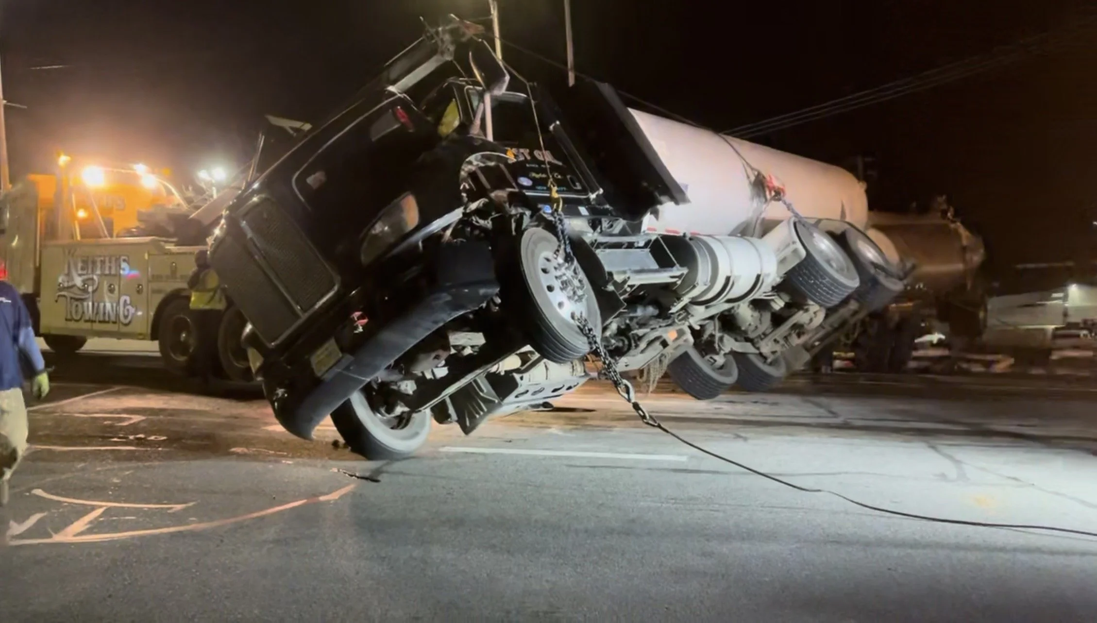 A large semi-truck is tipped onto its side on a dark road at night, with emergency and towing vehicles nearby.
