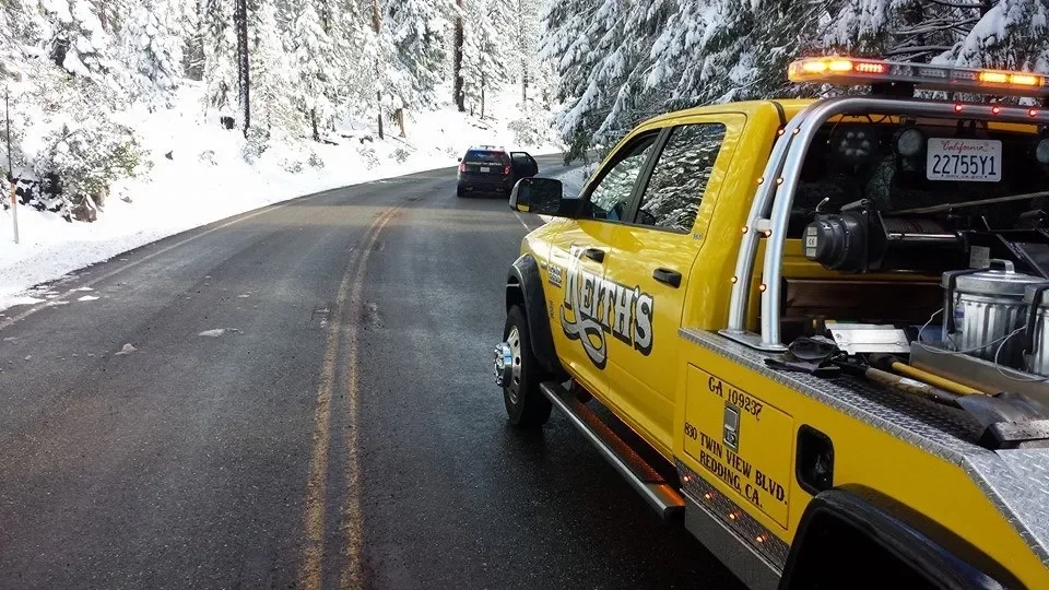 A yellow utility truck parked on a snowy mountain road with two cars ahead, surrounded by snow-covered trees.