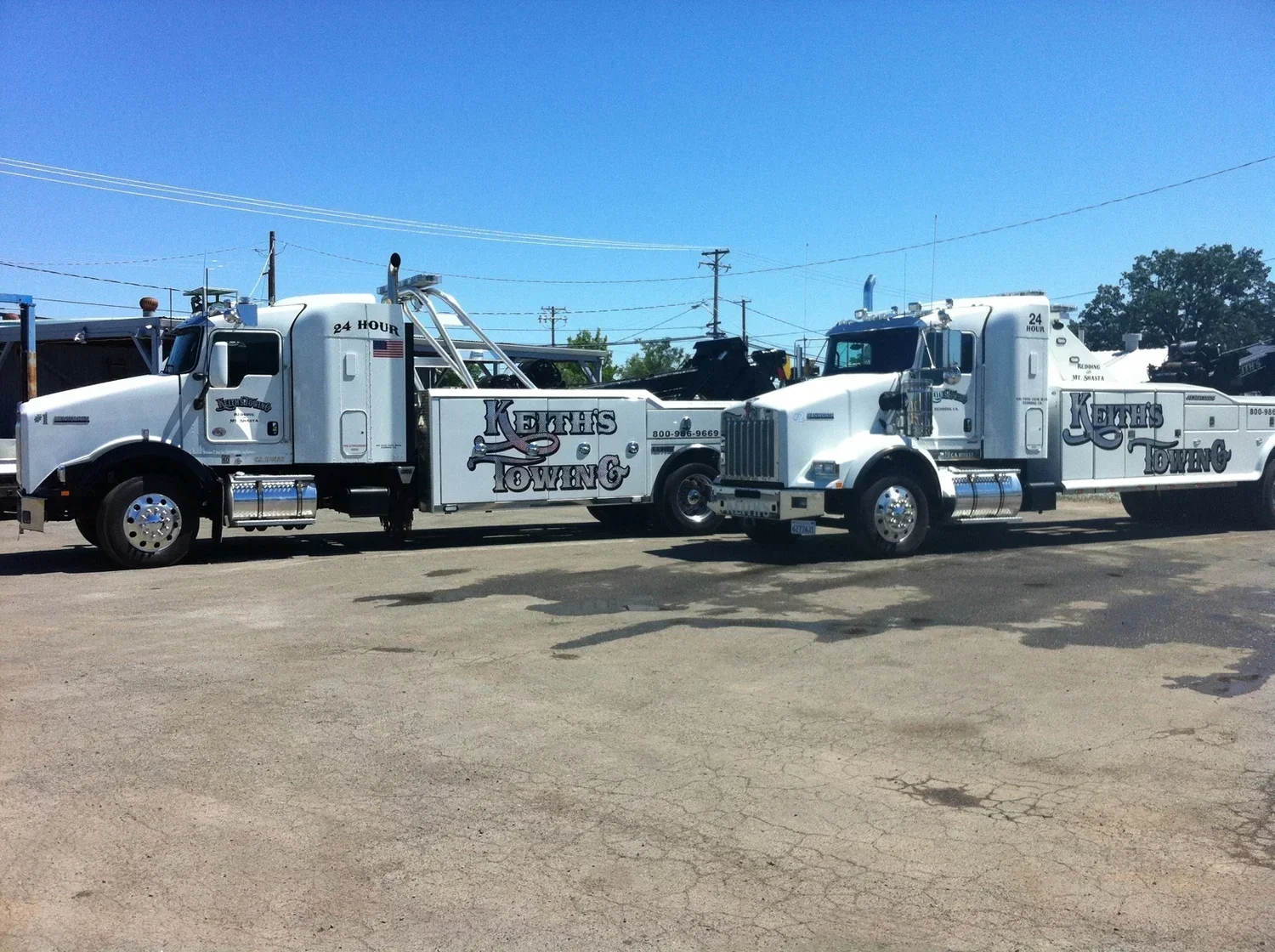Two white tow trucks with 'Keths Towing' written on the sides, parked on a concrete lot under a blue sky with power lines visible in the background.