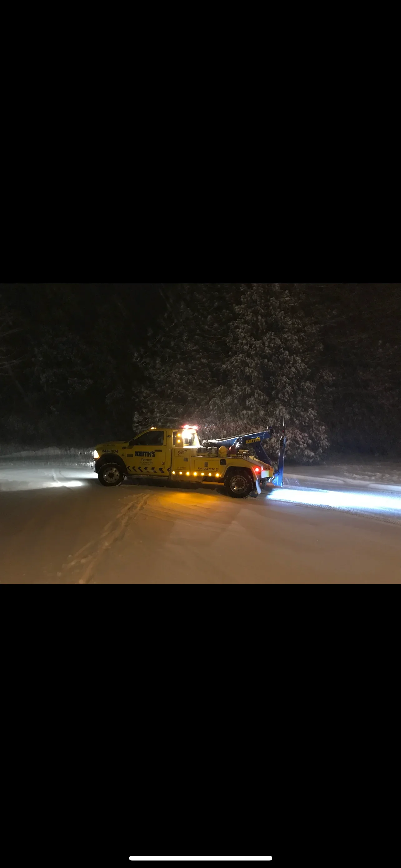 A yellow tow truck with flashing lights working in a snowy, dark night in a wooded area.