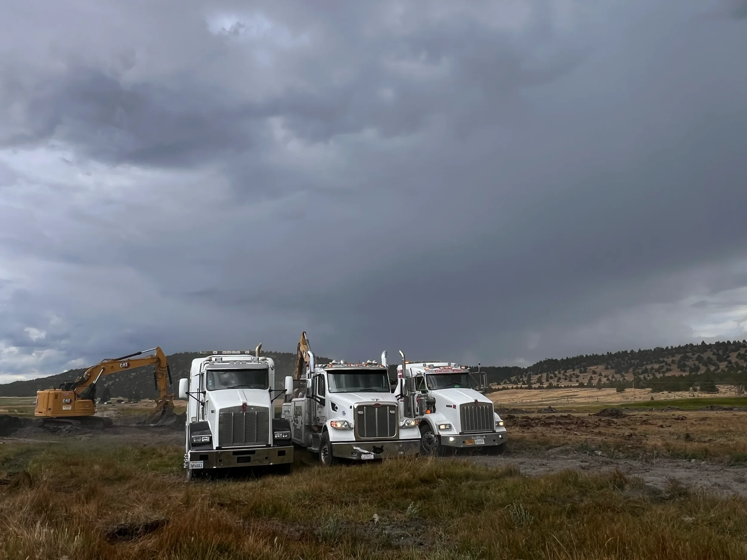 Three white semi-trucks and a yellow excavator working on a construction site under dark, cloudy sky with hills in the background.