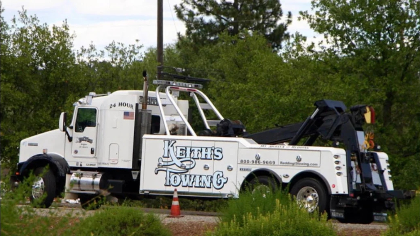 A white towing truck with 'Keith's Towing' written on the side, parked on a grassy area with green trees in the background.