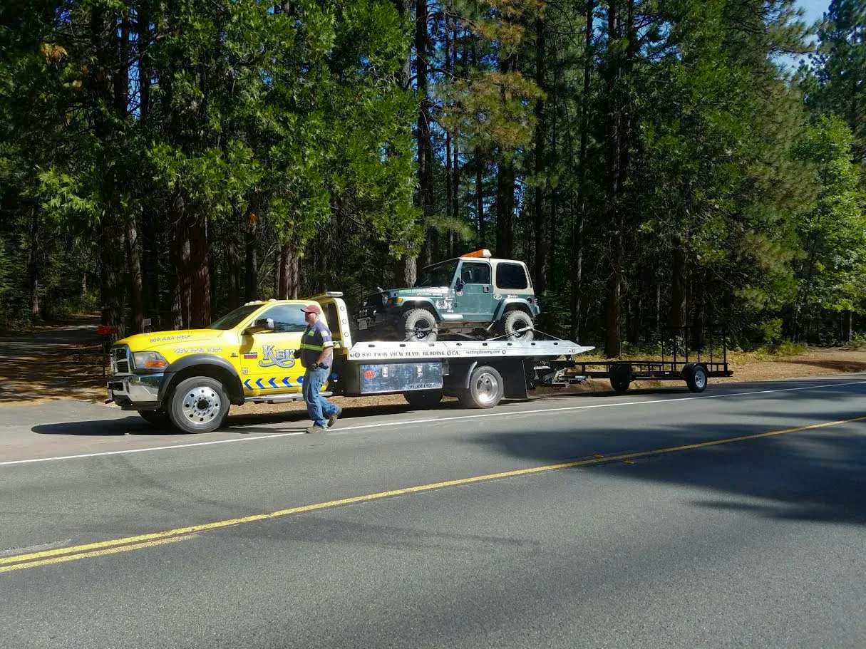 A tow truck on a road with a small off-road vehicle loaded on it, and a person walking nearby, with a forested area in the background.