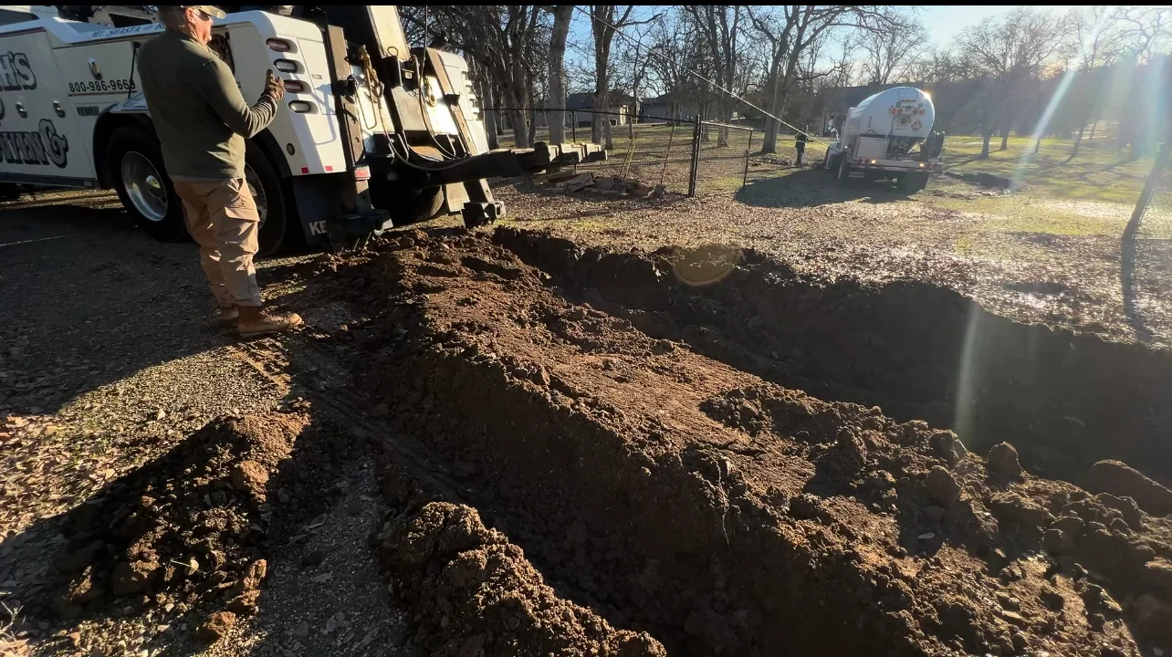 A worker standing next to a large bucket truck, digging a trench in a ground area, with dirt piled up beside the excavation; there is a water truck in the background.