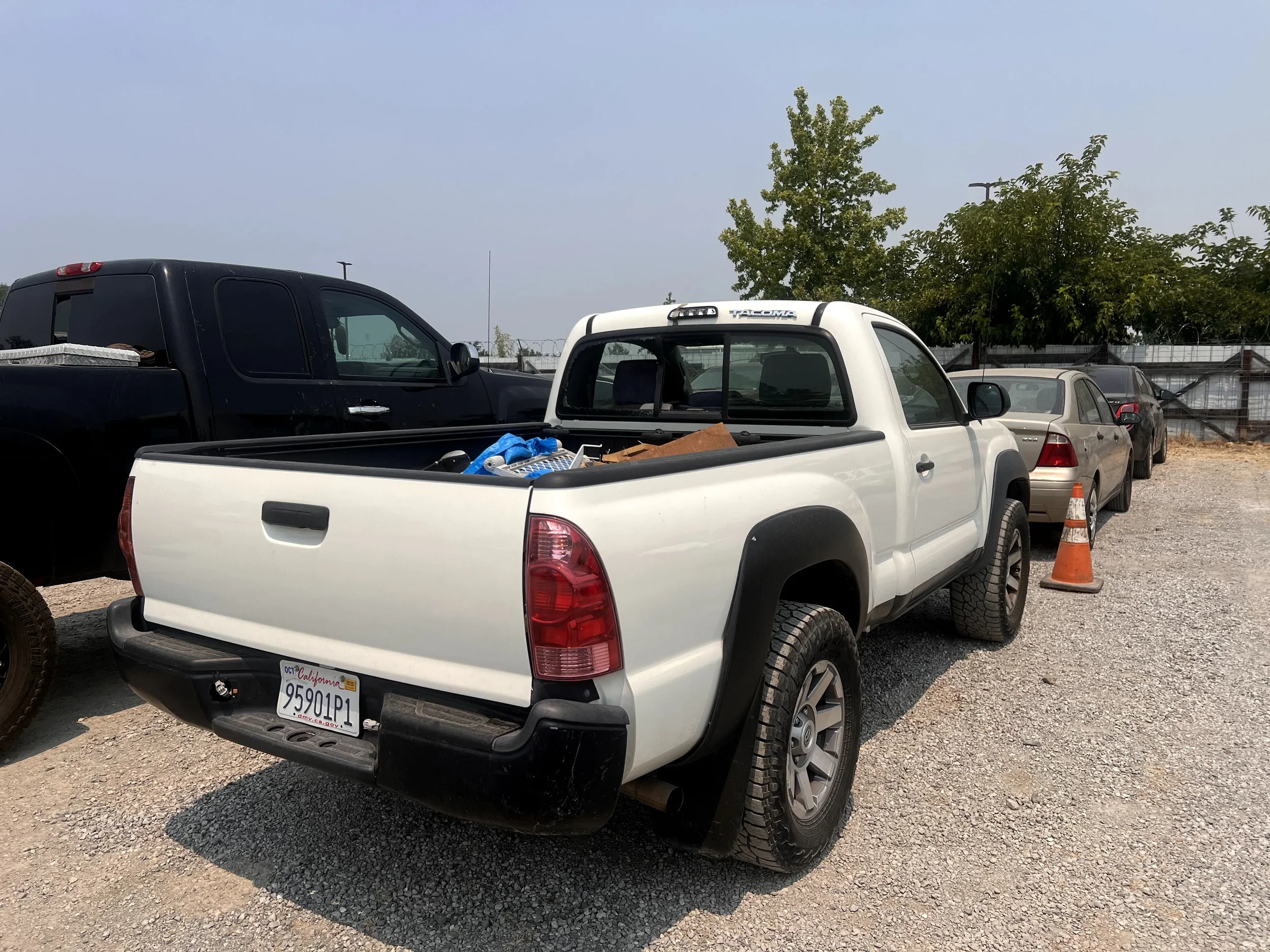 White Toyota Tacoma pickup truck parked in a lot with other vehicles, a traffic cone, and trees in the background.