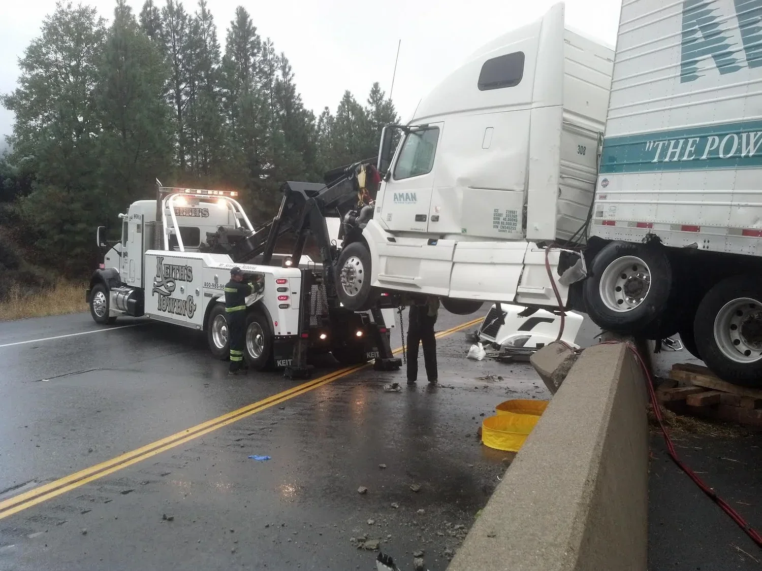 Semi-truck involved in a crash with a concrete barrier on a wet road, emergency responders at the scene.