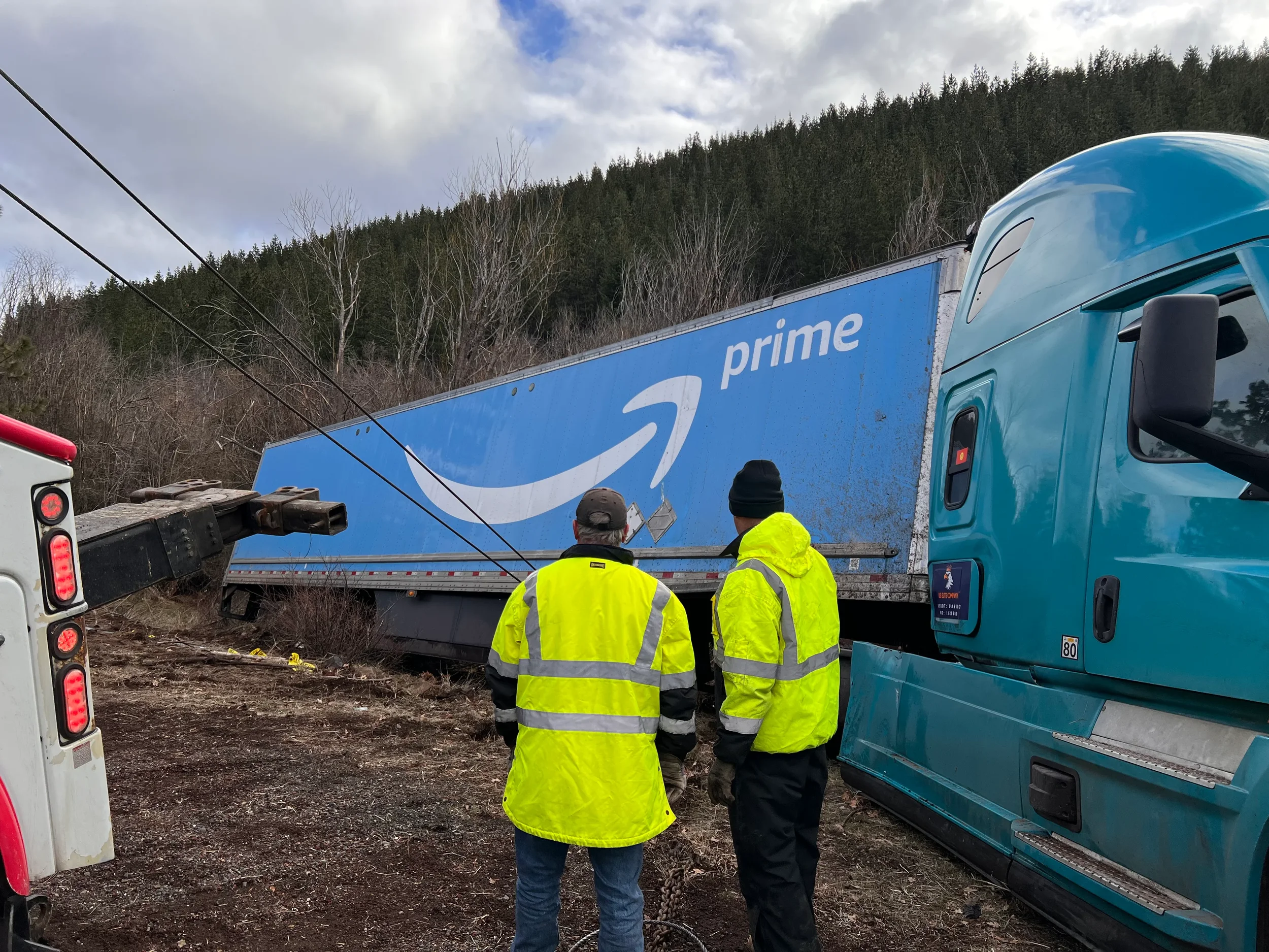 Two workers in yellow safety jackets stand in front of a Semi-truck with an Amazon Prime trailer that has collided and is tilted on the side of a dirt road. A crane is nearby, possibly involved in the accident.
