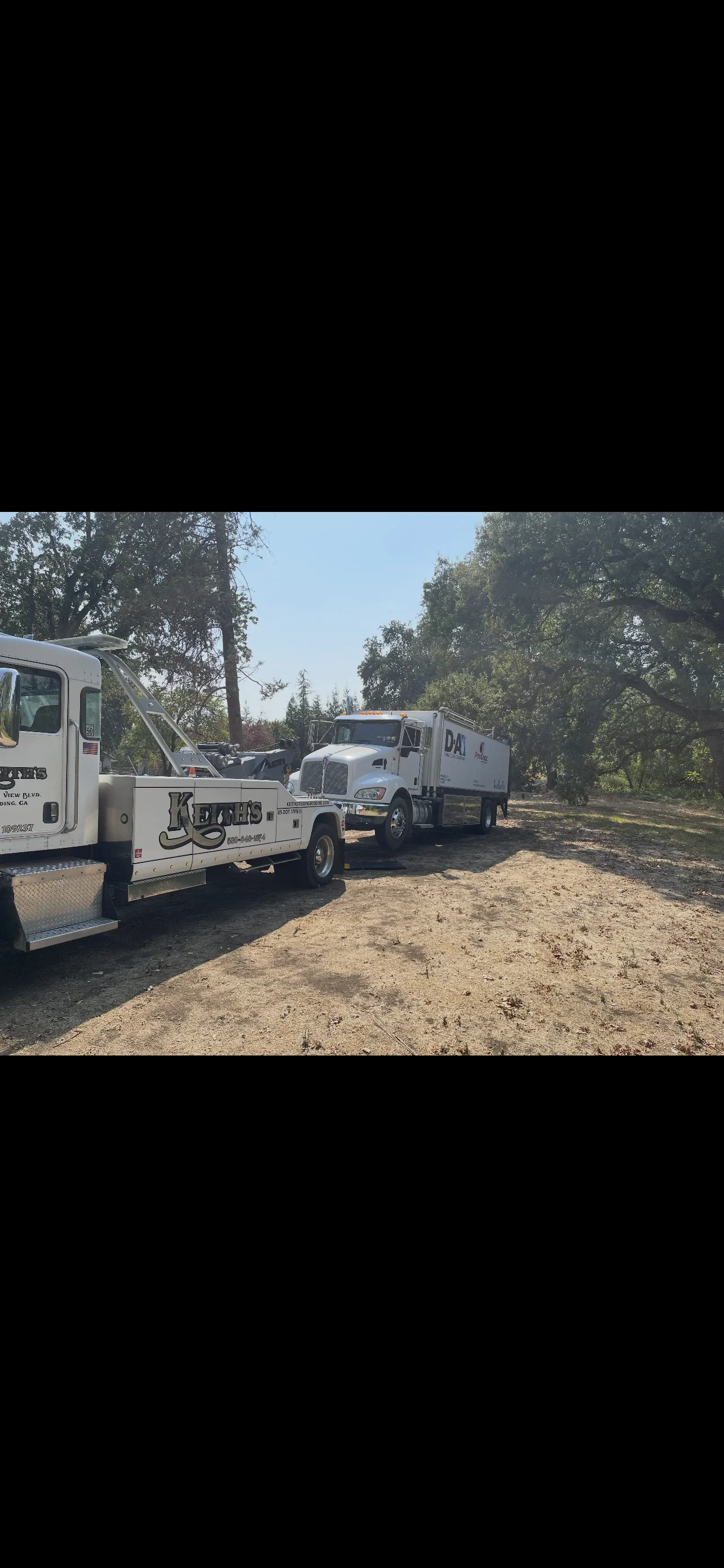 Two tow trucks parked on a dirt area with trees around, one with a flatbed and the other with a box truck.