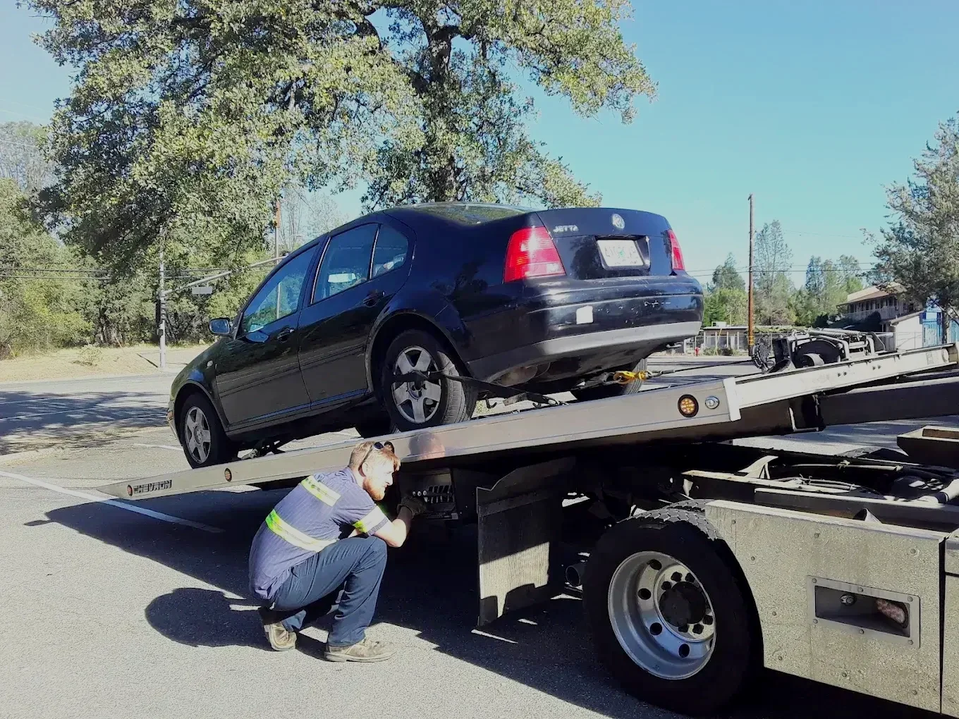 A tow truck transporting a black Volkswagen rear-ended vehicle on a parkway during daytime, with a firefighter inspecting the vehicle.