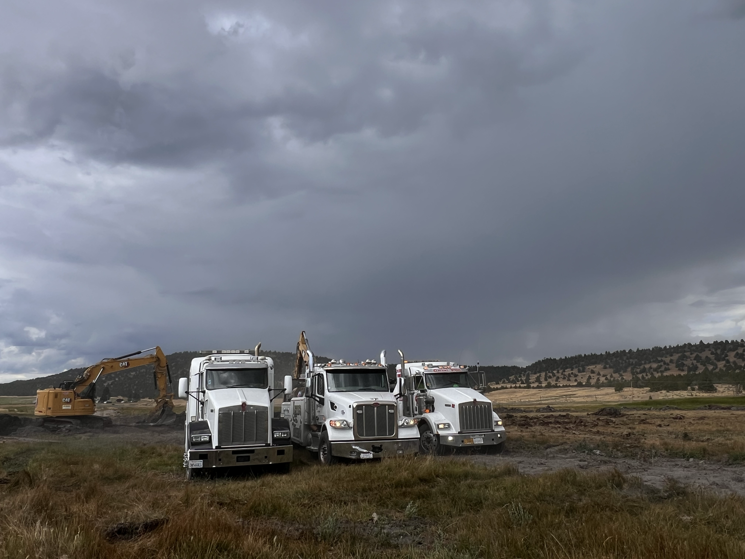Three large white semi-trucks parked on a grassy field under a dark, cloudy sky, with a yellow excavator in the background.