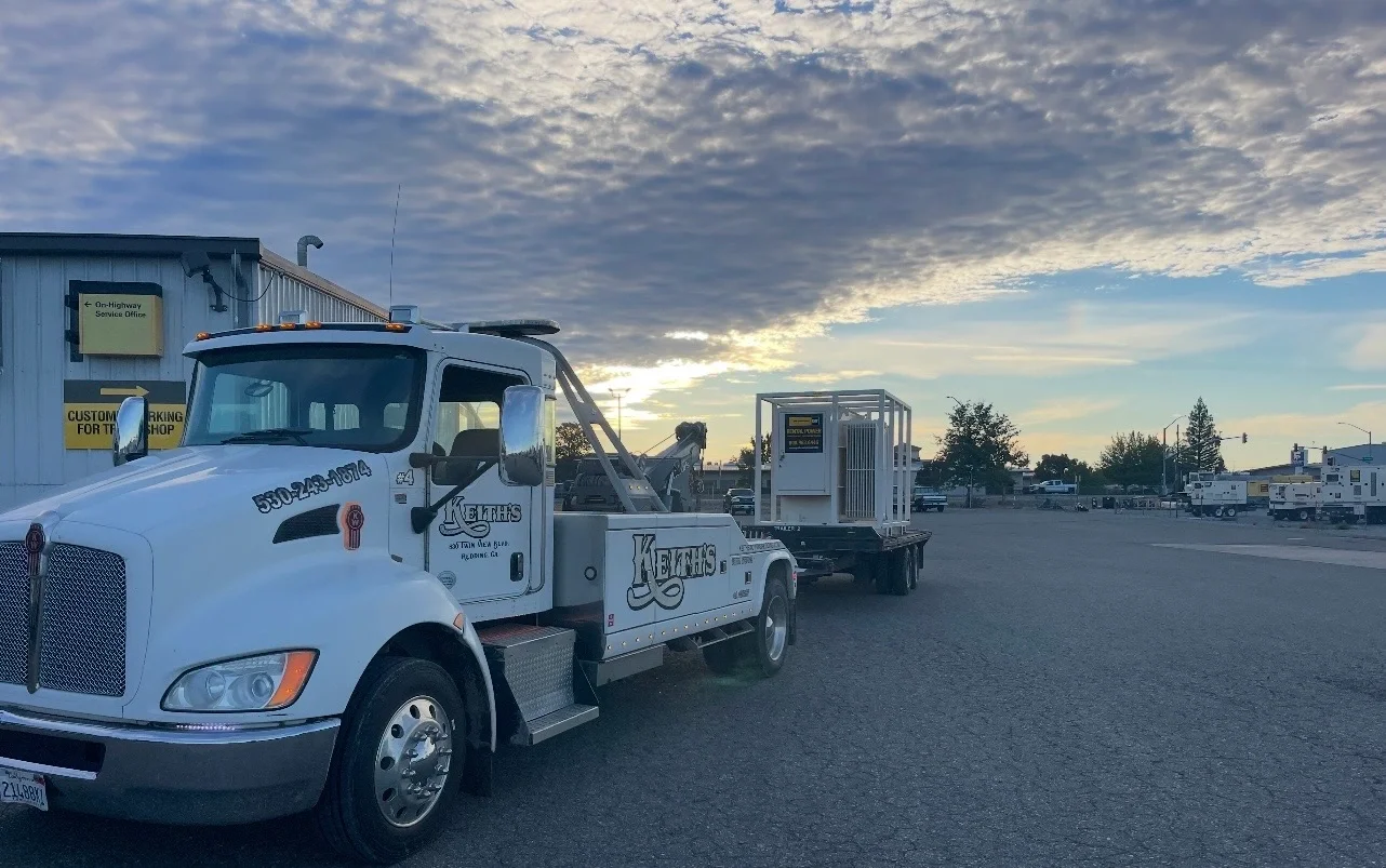 A white tow truck with equipment on its bed, parked in a lot near a building with a sky filled with clouds during sunset.