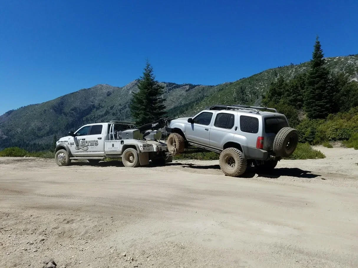 A tow truck is towing a gray SUV on a dirt road in a mountainous area with trees and blue sky.