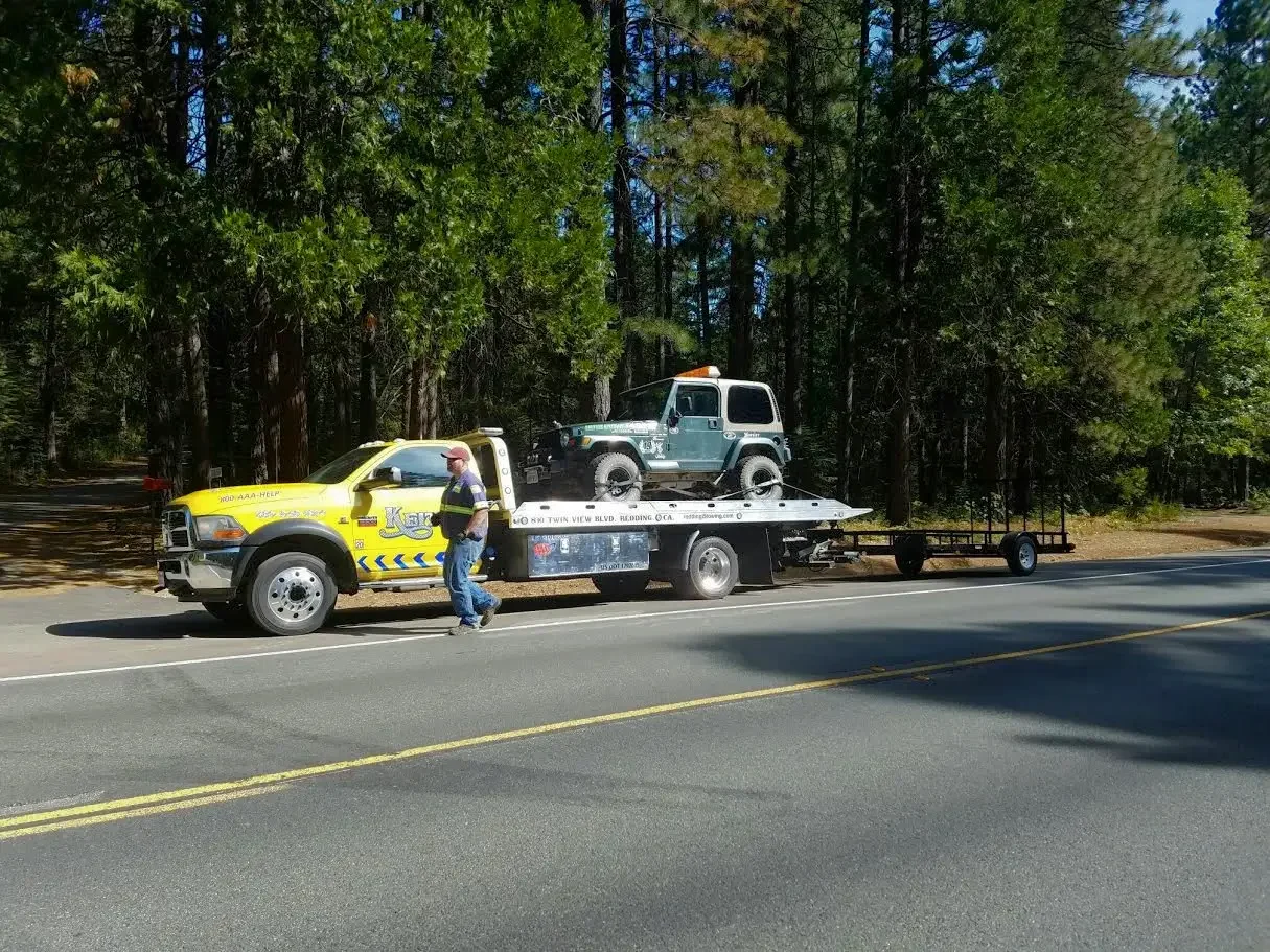 A tow truck carrying a small green off-road vehicle on a rural road with trees in the background.