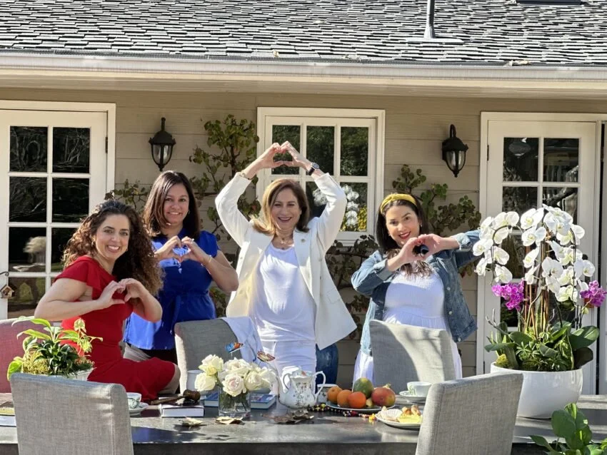 Four women standing outdoors at a table, smiling and making heart shapes with their hands, with a house in the background, flowers on the table, and bright sunlight.