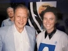 A woman with short blonde hair and an older man with gray hair smiling together, with two people blurred in the background.