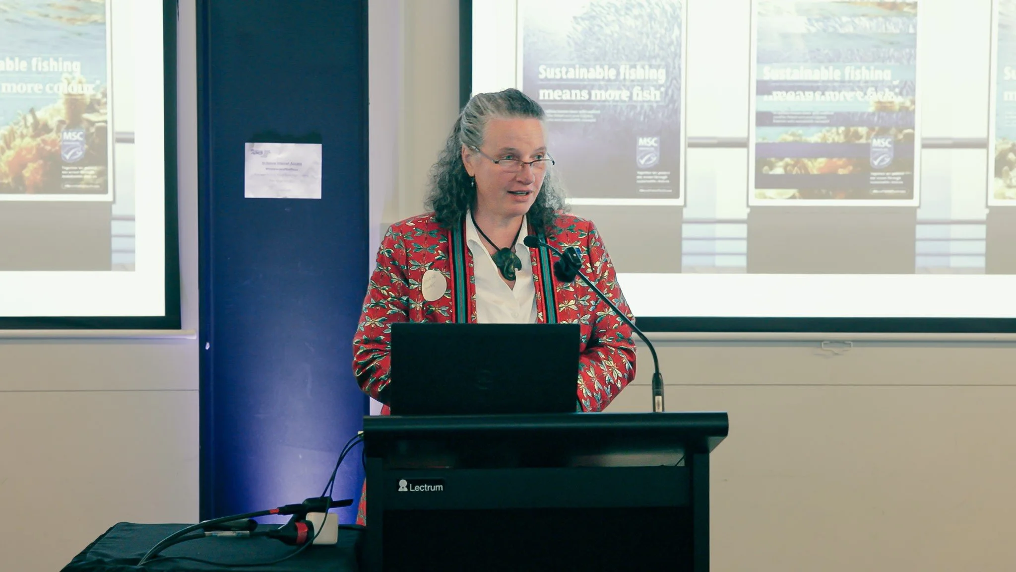 Woman giving a presentation at a podium with a laptop, wearing glasses and a red patterned blazer, with posters about sustainable fishing behind her.