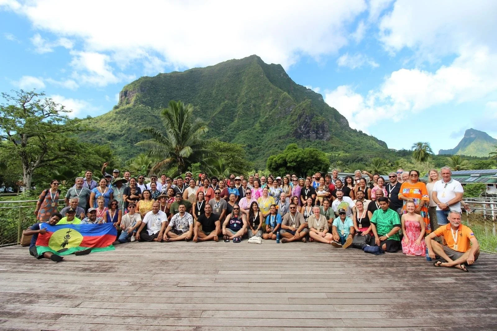 A large group of diverse people posing for a photo outdoors on a wooden platform, with lush green mountains and blue sky in the background.