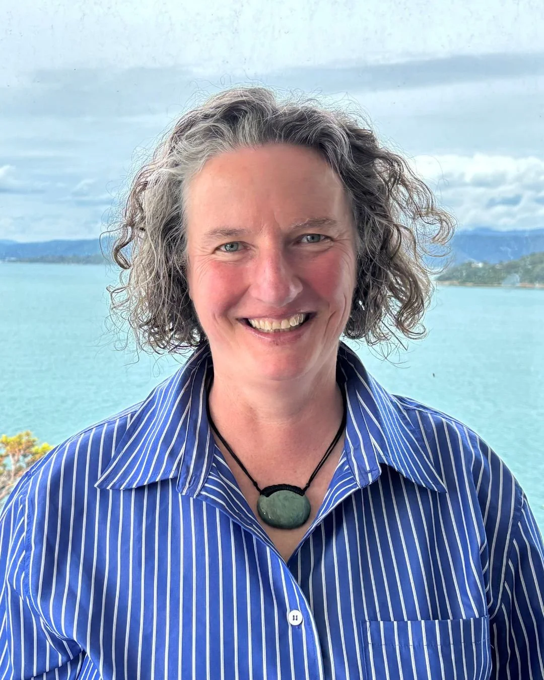 A woman with short curly grey hair smiling, wearing a blue and white striped shirt and a green pendant necklace, with a body of water and distant hills in the background.