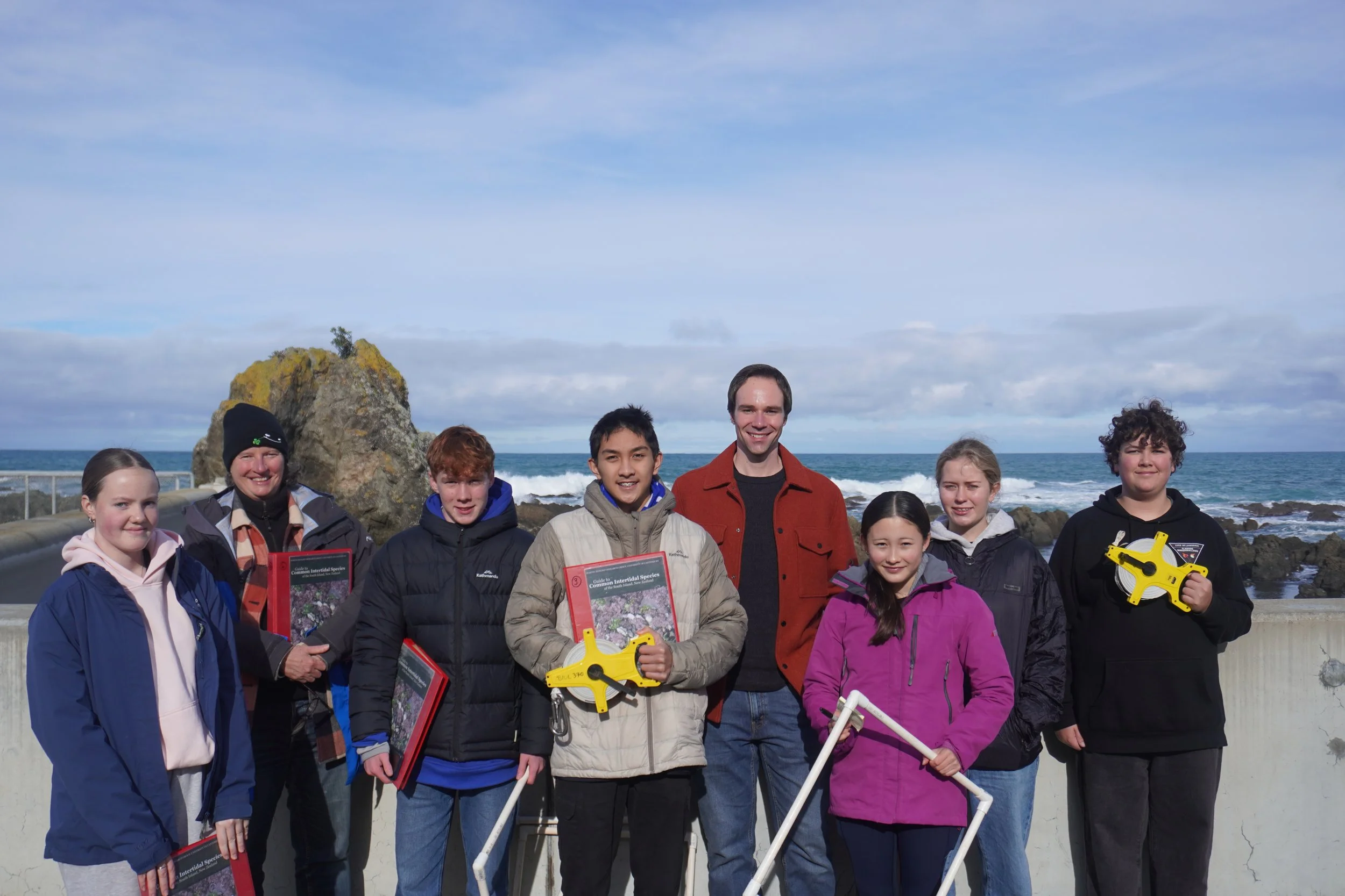 Group of people standing on a beach near the ocean with rocks and a cloudy sky in the background. They are holding books and tools.