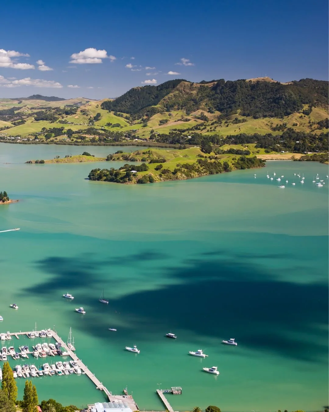 Aerial view of a turquoise bay with boats, green hills, and distant mountains under a blue sky with white clouds.