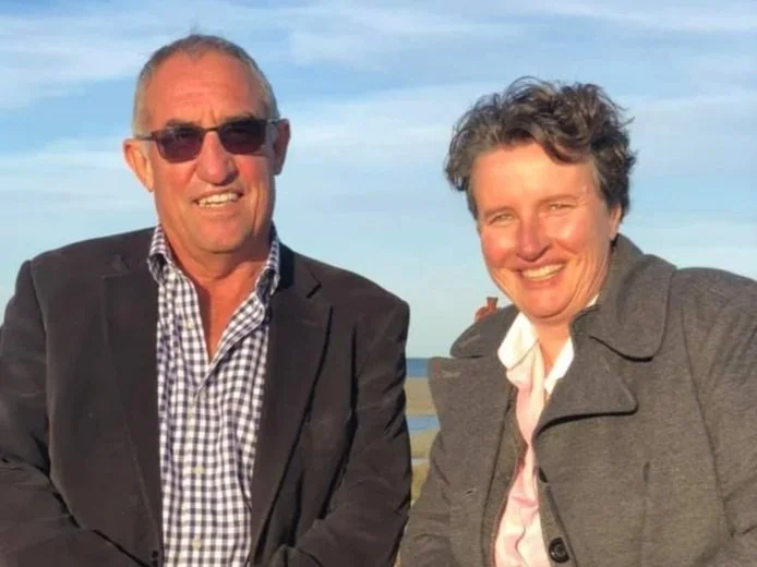 A man and a woman smiling outdoors with a body of water in the background, dressed in smart casual clothing.