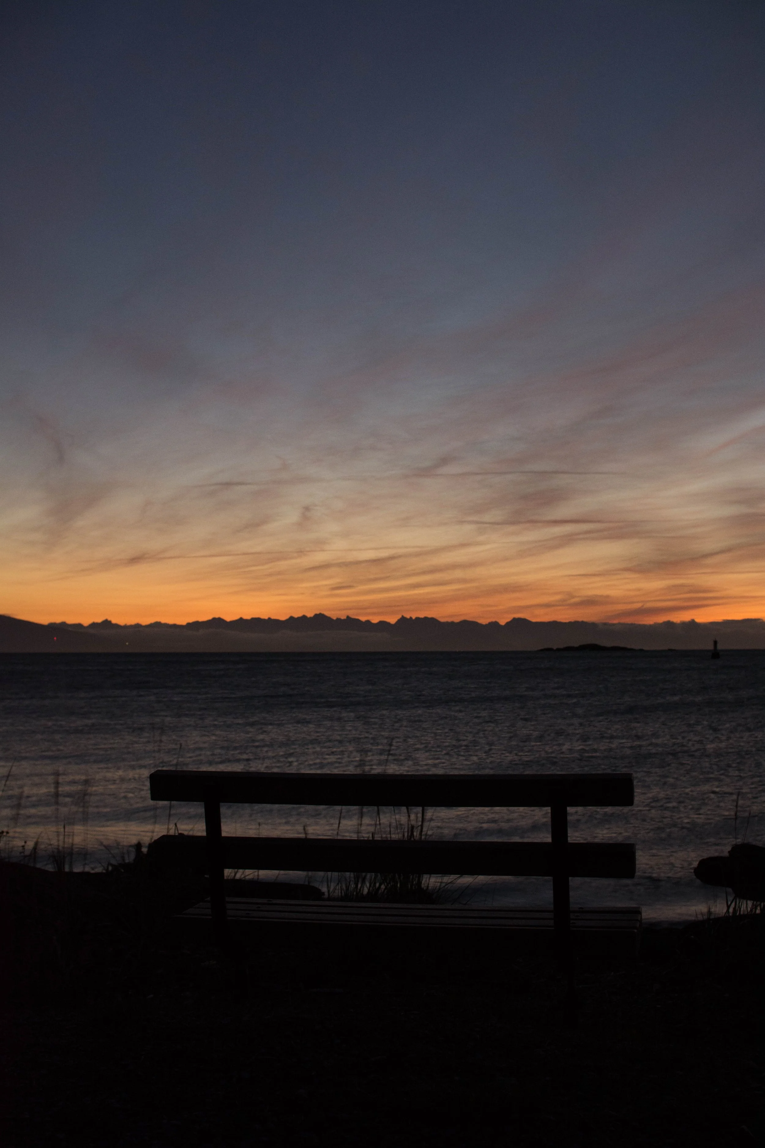 A bench facing a body of water during sunset with a colorful sky and mountains in the distance.