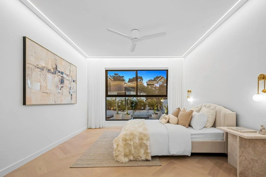 Bright, minimalist bedroom with white walls, large window, bed with white bedding and beige cushions, and a textured nightstand.