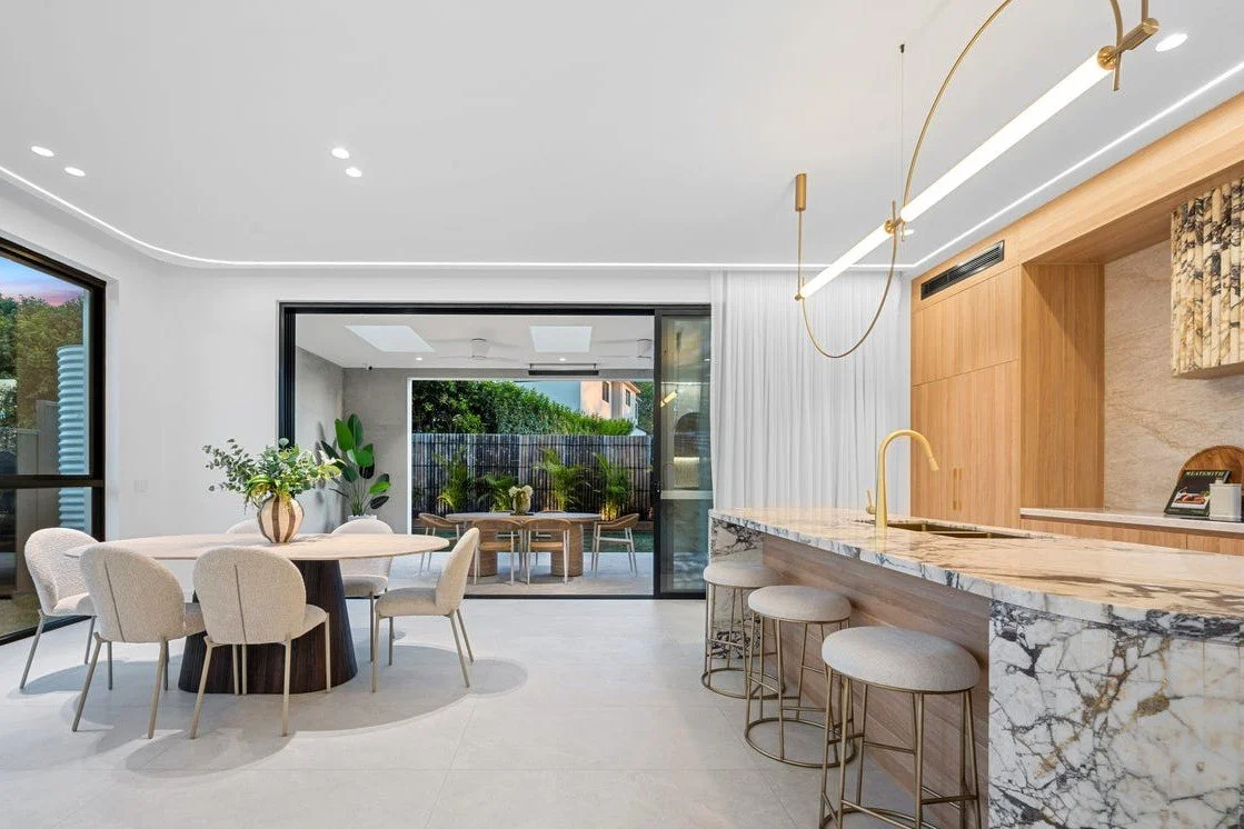 Modern open-concept kitchen and dining area with a marble island, gold bar stools, and a round dining table with beige chairs. Glass sliding doors lead to an outdoor patio with greenery, and the space is decorated with a vase of flowers on the table.