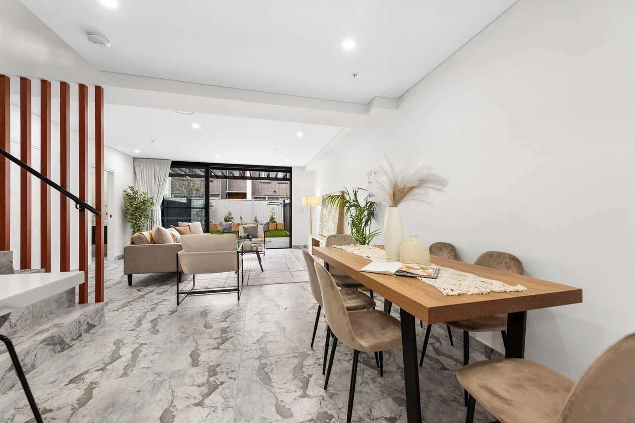Modern open-concept living and dining area with marble floors, a wooden dining table with six chairs, a sofa set facing a sliding glass door, and a small outdoor patio with plants.