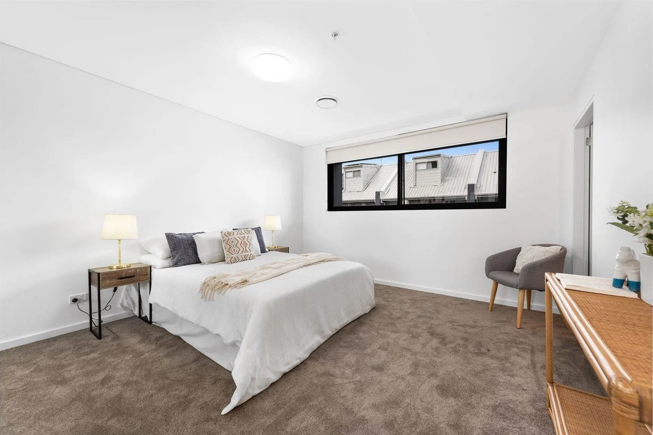 A bright, minimalist bedroom with a large bed, white bedding, and pillows, bedside lamps, a window with a view of rooftops, a gray armchair, and a wooden dresser with a plant and decorative items.