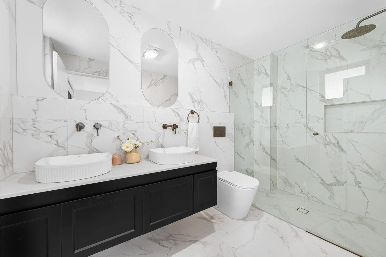 Modern bathroom with white marble walls and floor, black vanity with two white vessel sinks, wall-mounted faucets, oval mirrors, toilet, and glass-enclosed shower with rainfall showerhead.
