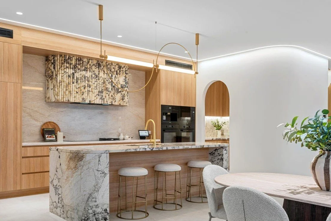 Modern kitchen with wooden cabinets, marble island, built-in black appliances, and a marble backsplash. There is a decorative curtain above the stove, gold pendant lights, and a dining area with a wooden table and gray upholstered chairs.