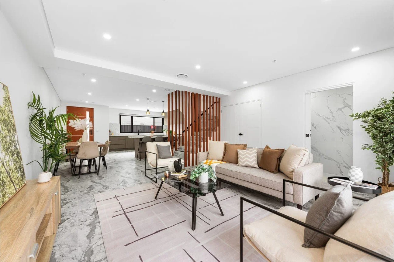 Spacious living room with white walls and marble flooring, beige sofas with cushions, a glass coffee table with decorative items, a pink rug, a dining table with beige chairs, a wooden partition with stairs, plants, and a modern kitchen in the background.