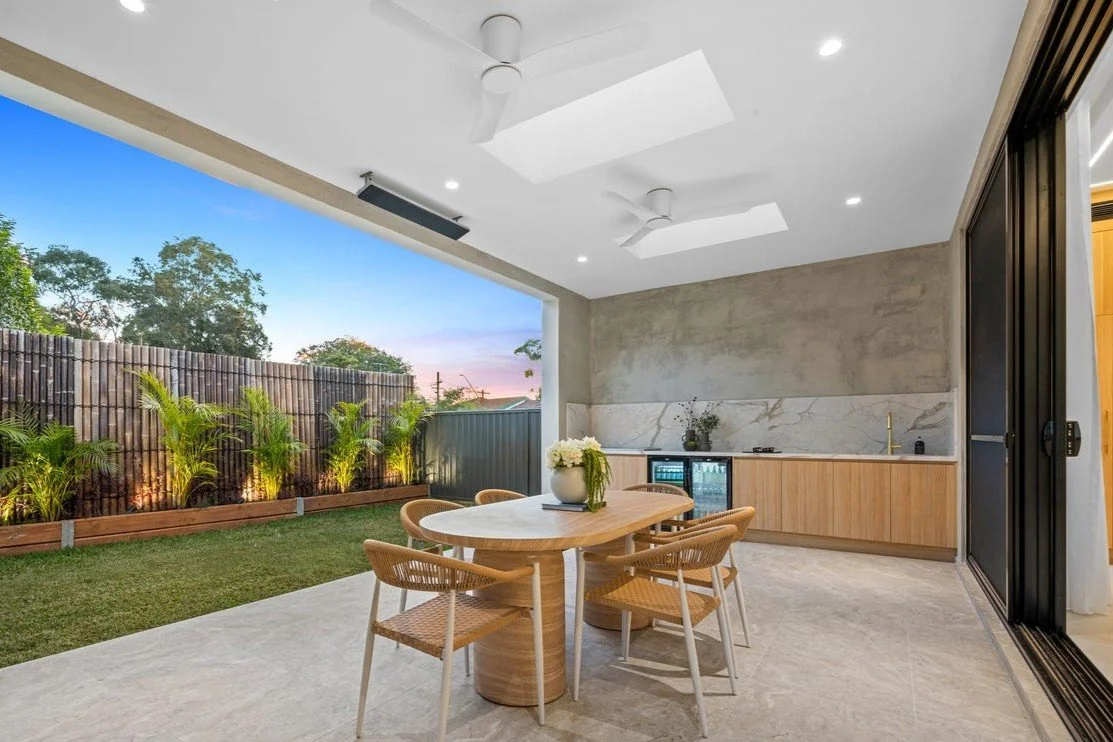 Modern outdoor patio with a dining table and chairs, overlooking a backyard with green plants and a wooden fence, under a sky at sunset.