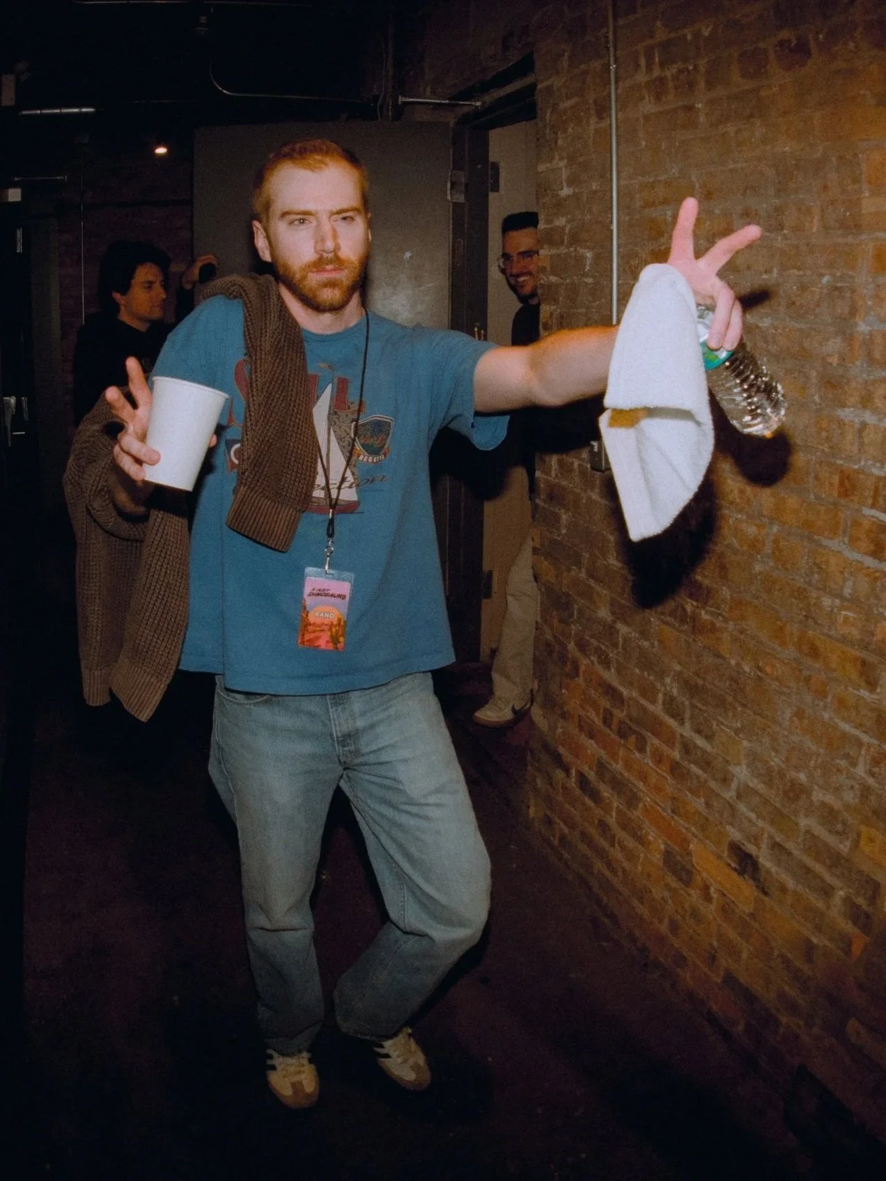 A man with reddish hair and a beard posing indoors against a brick wall, making a peace sign with his right hand. He is holding a water bottle and a cup in his left hand, has a brown jacket draped over his shoulder, and wears a blue T-shirt with a graphic design, jeans, and sneakers. There are other people in the background.