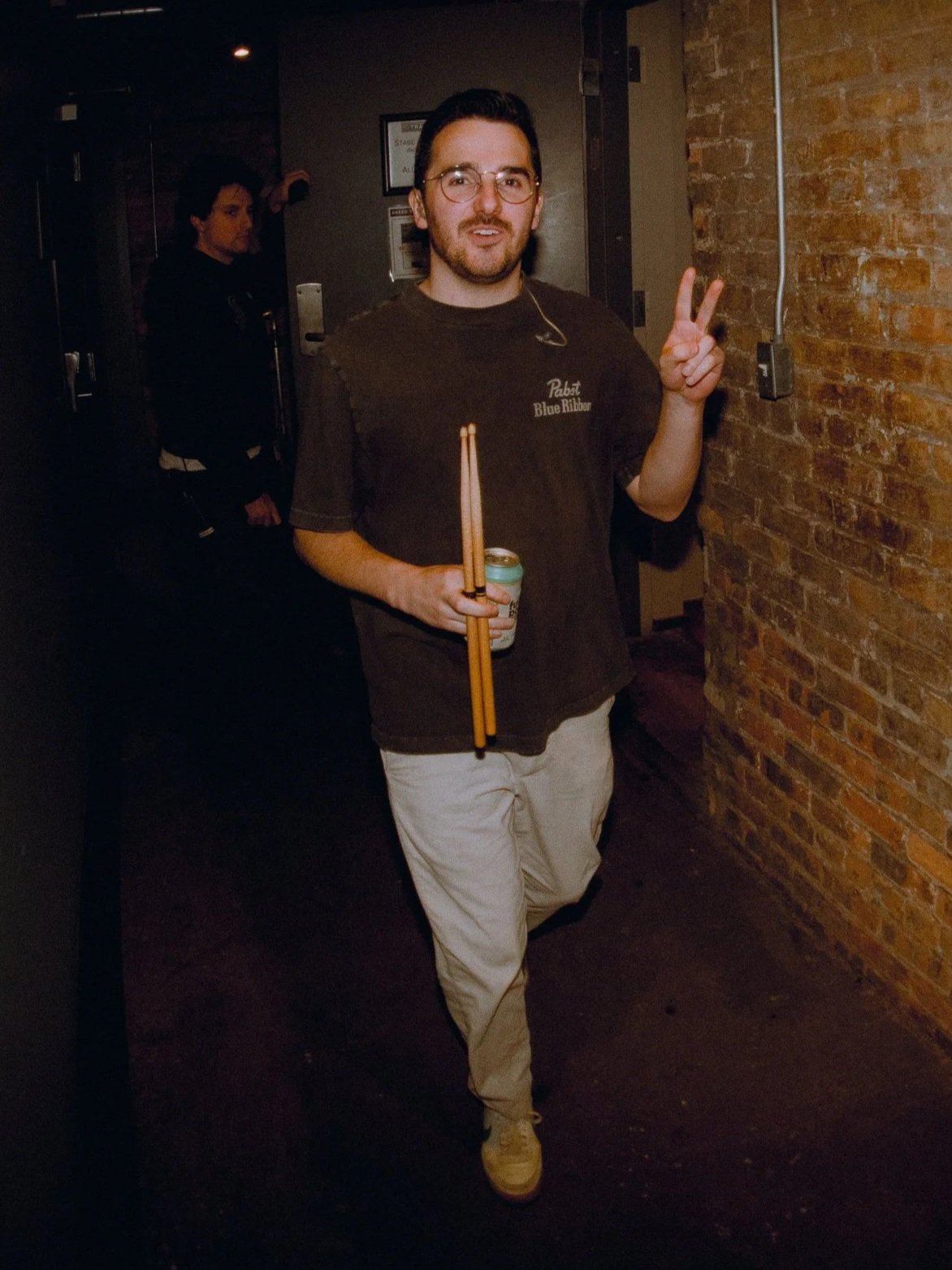 A man wearing glasses and a brown T-shirt with 'Pabst Blue Ribbon' logo, holding a drink and drumsticks, making a peace sign with his right hand, standing in a dimly lit room with a brick wall and another person in the background.