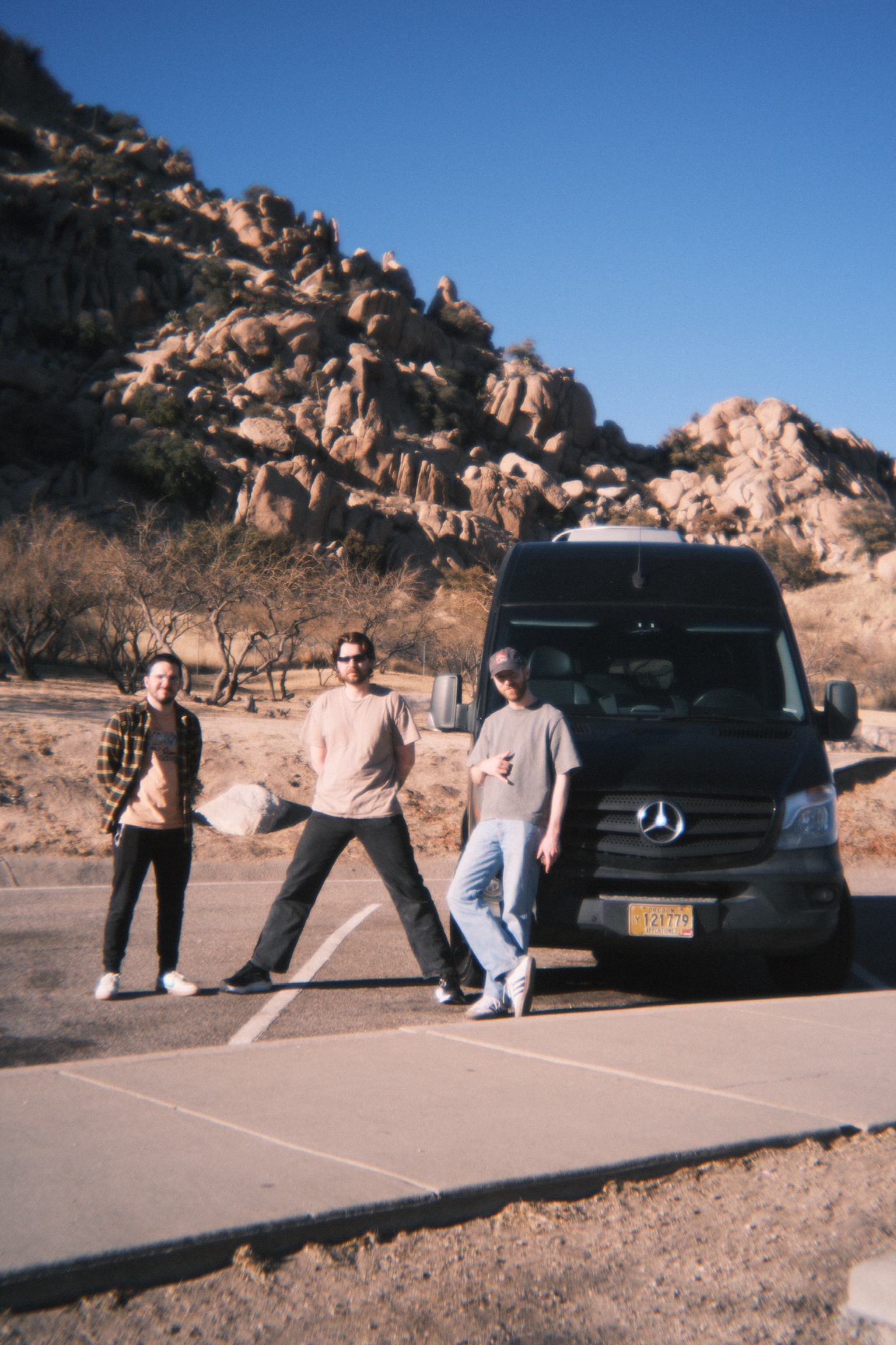 Three young men standing in front of a black Mercedes-Benz van in a desert landscape with rocky hills and sparse vegetation, under a clear blue sky.