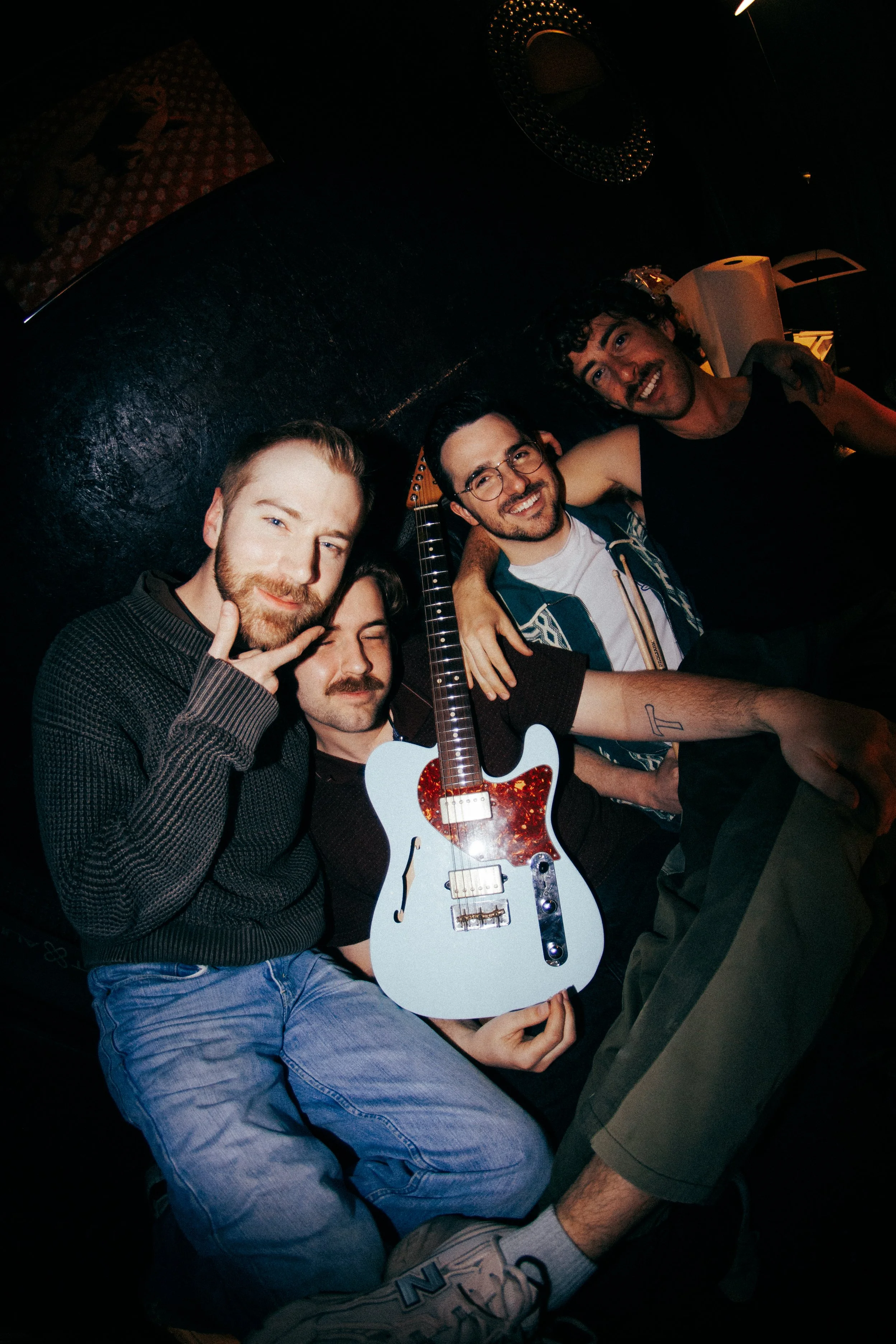 Group of five friends in a dimly lit room, smiling and posing for a photo with one holding a blue and red guitar.