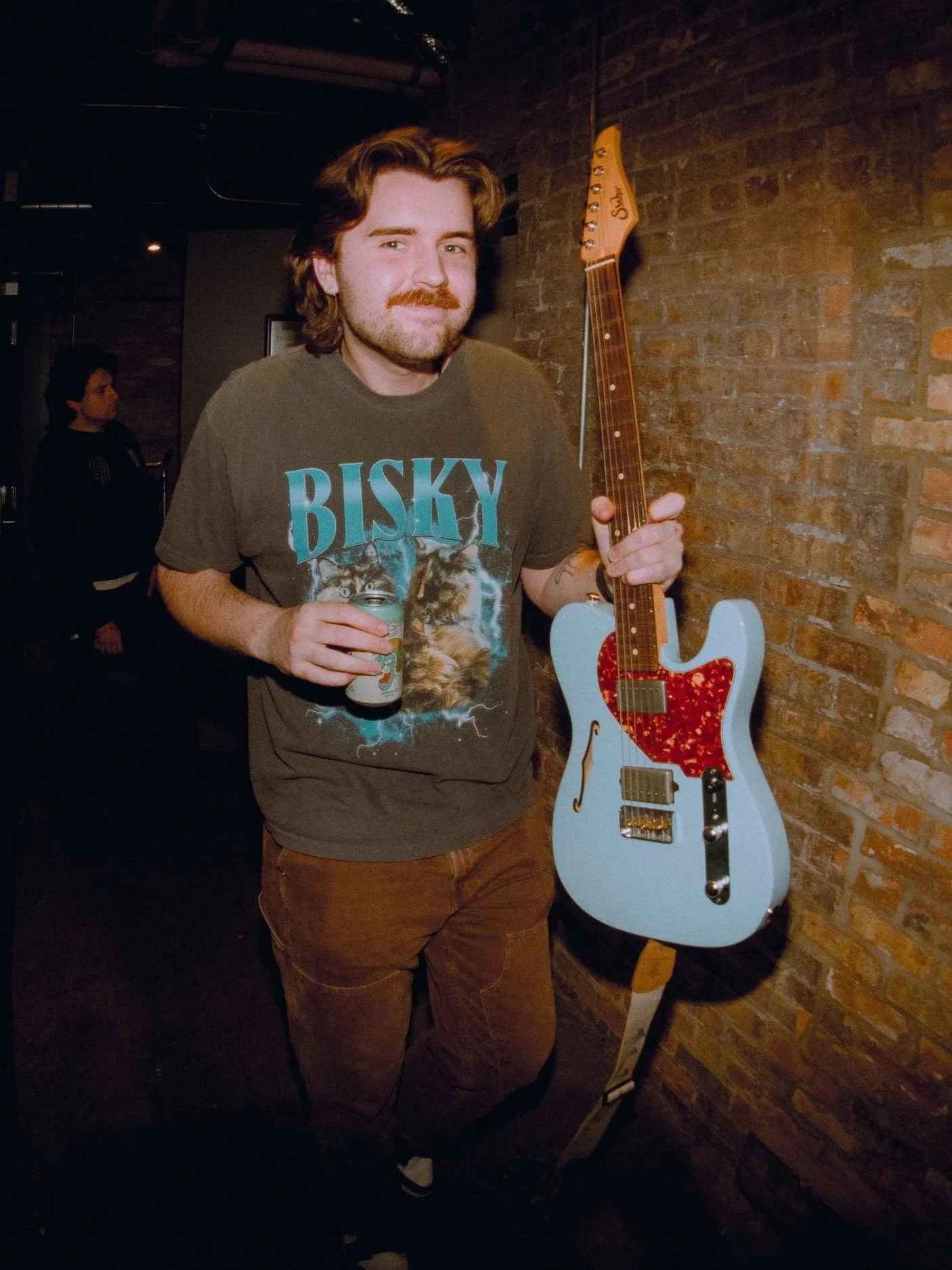 A man with shoulder-length brown hair and a beard holding a can of Bud Light beer, standing next to a white electric guitar with a red tortoiseshell pickguard, against a brick wall background.