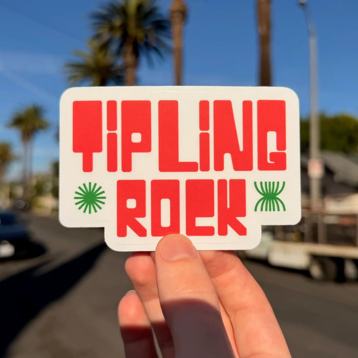 A hand holding a sticker that says "Tip Line Rock" in red, with green decorative elements, against a background of palm trees and a blue sky.