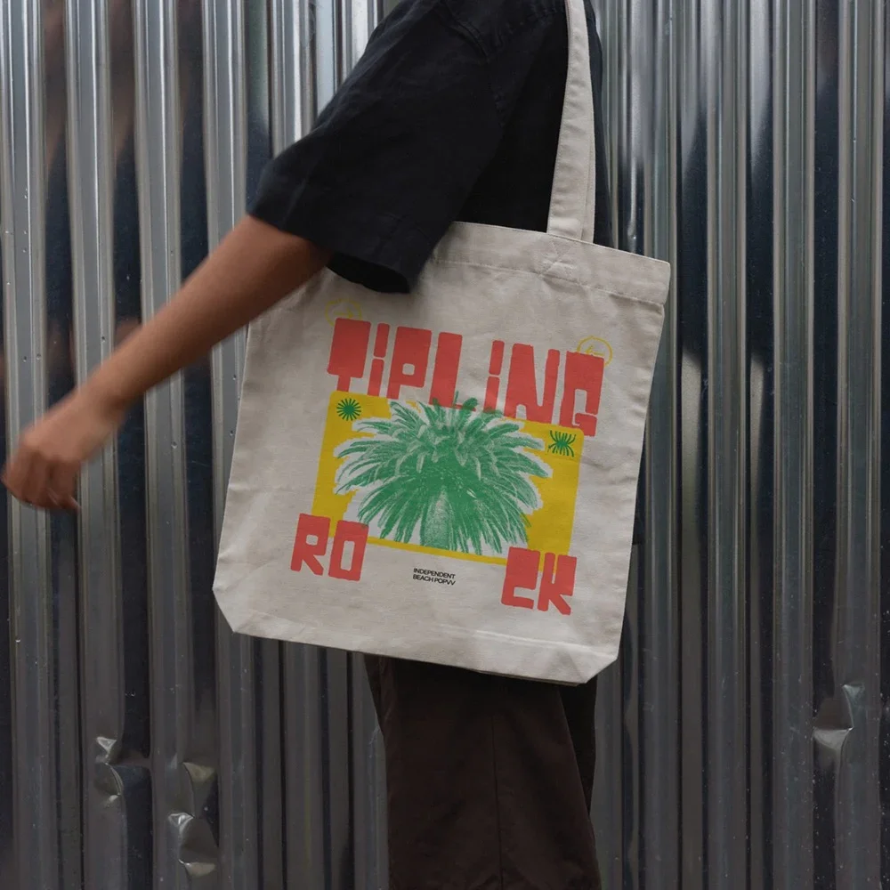 Person carrying a canvas tote bag with a palm tree graphic and text 'TIP JUNG RO' in front of metallic wall panels.
