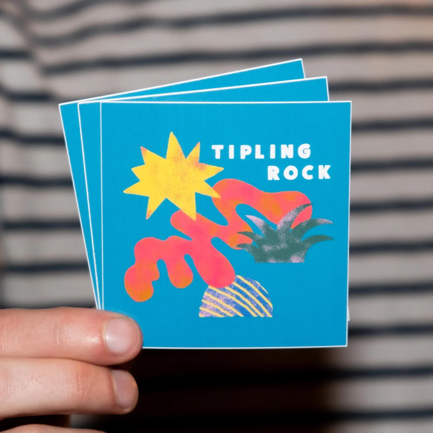 A hand holding stickers with the name 'Tipling Rock' and an abstract illustration.
