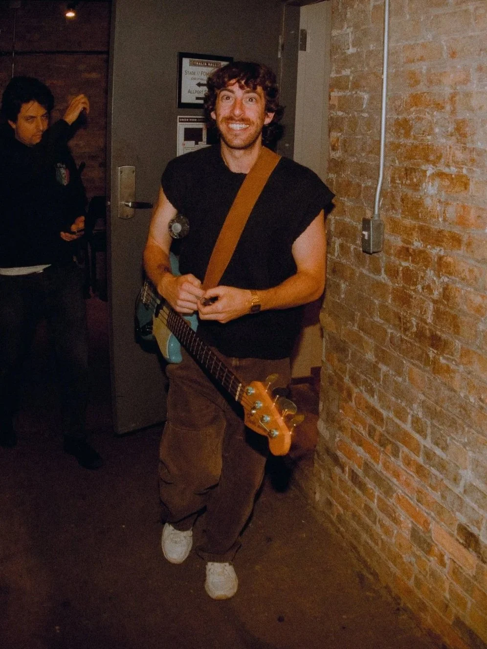 A man with curly hair and a beard, smiling, holding a guitar, standing in a brick-walled room.