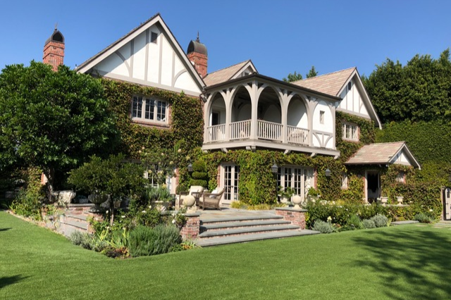 Front view of a large, white, Tudor-style house with a garden and lawn, surrounded by greenery and trees.