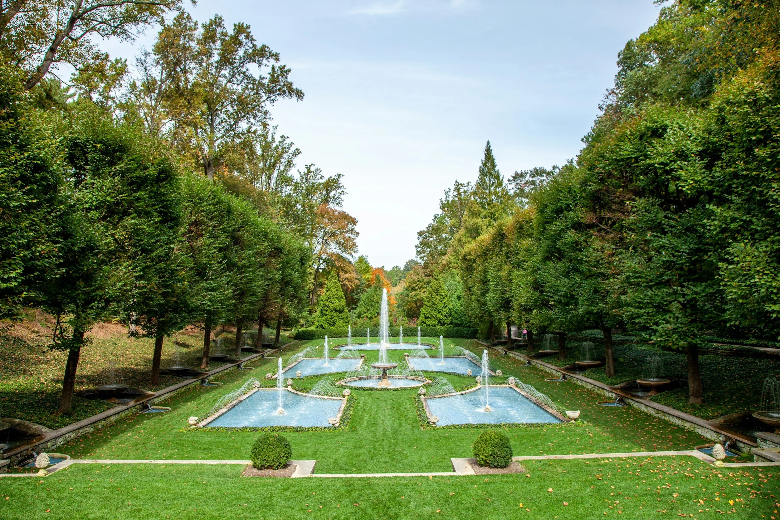 Landscape view of a large park with a fountain in the center, surrounded by neatly trimmed grass, small bushes, and tall trees with autumn foliage.